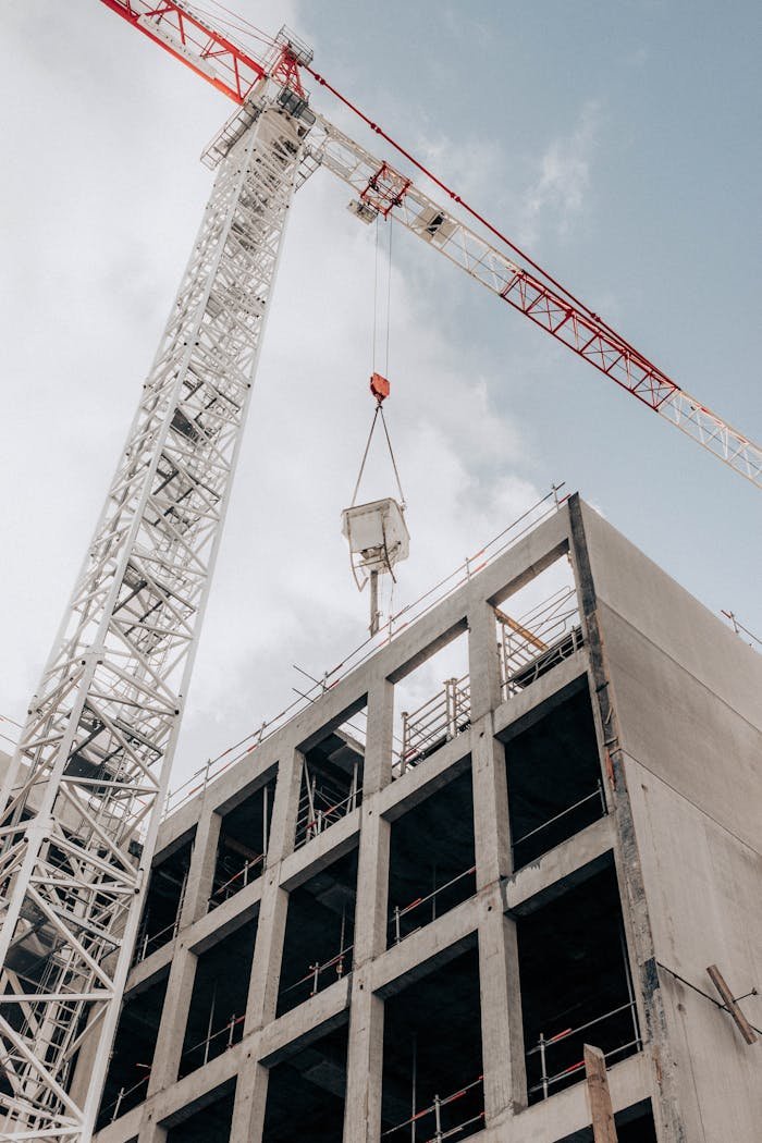 From below of modern hydraulic luffing jib tower crane working at construction site near modern unfinished multistory building under cloudy sky in city